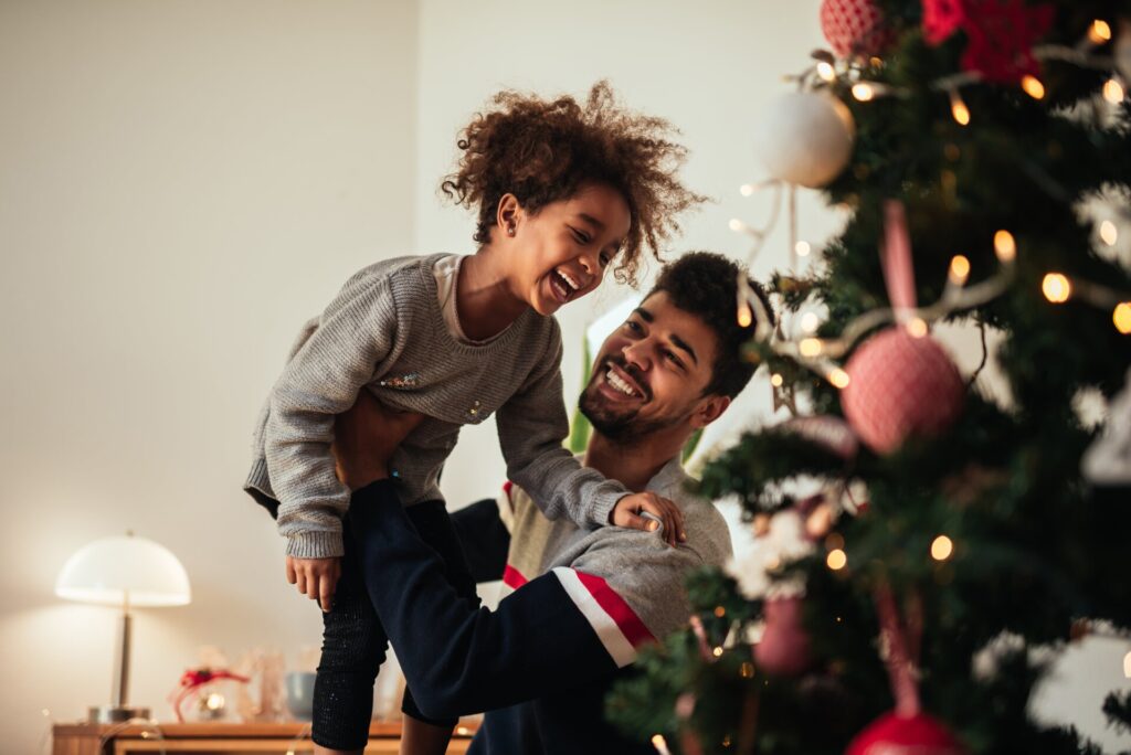 A man lifts a laughing child in front of a beautifully decorated Christmas tree, capturing the joy and warmth of the holiday season.