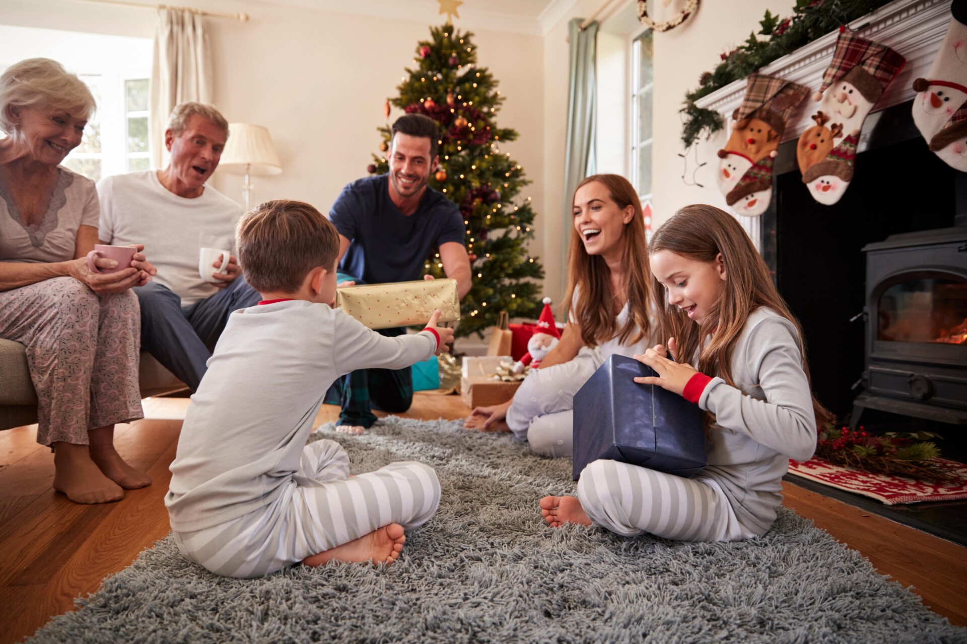 Family exchanging gifts in a cozy living room with a Christmas tree and stockings. Two children in pajamas sit on the floor, surrounded by adults.