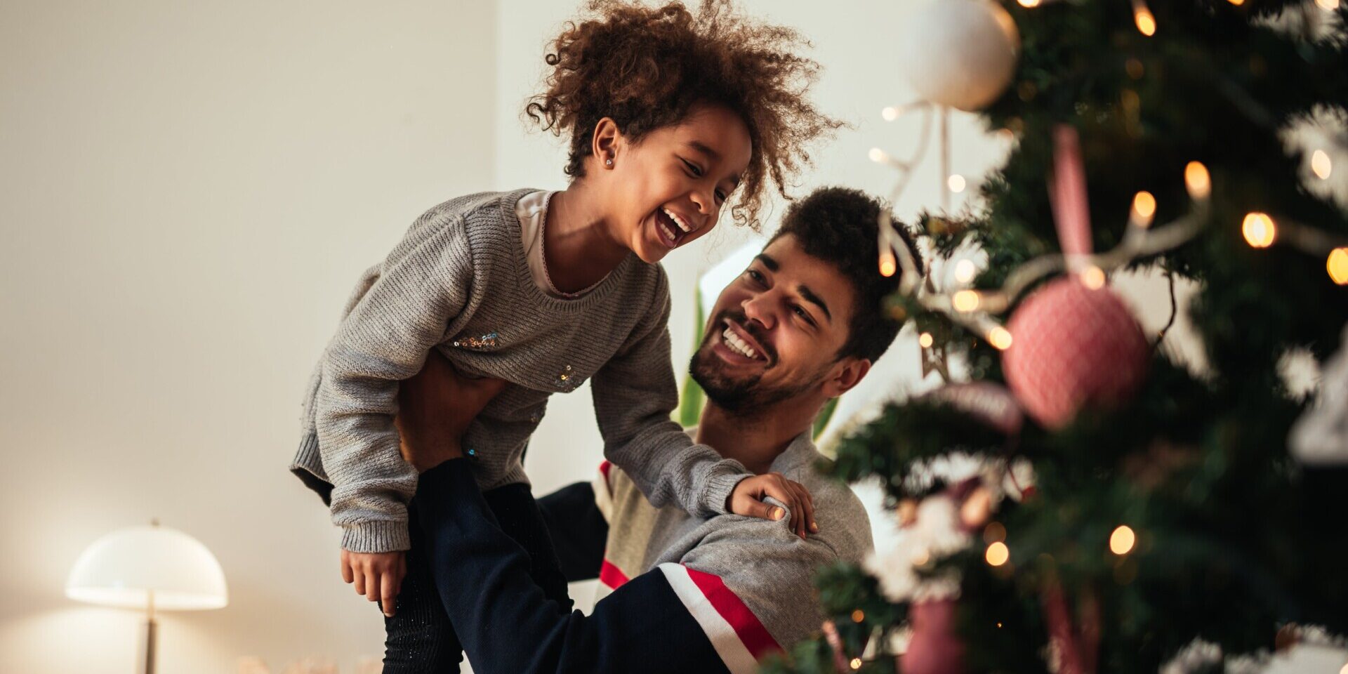 A man lifts a laughing child in front of a beautifully decorated Christmas tree, capturing the joy and warmth of the holiday season.