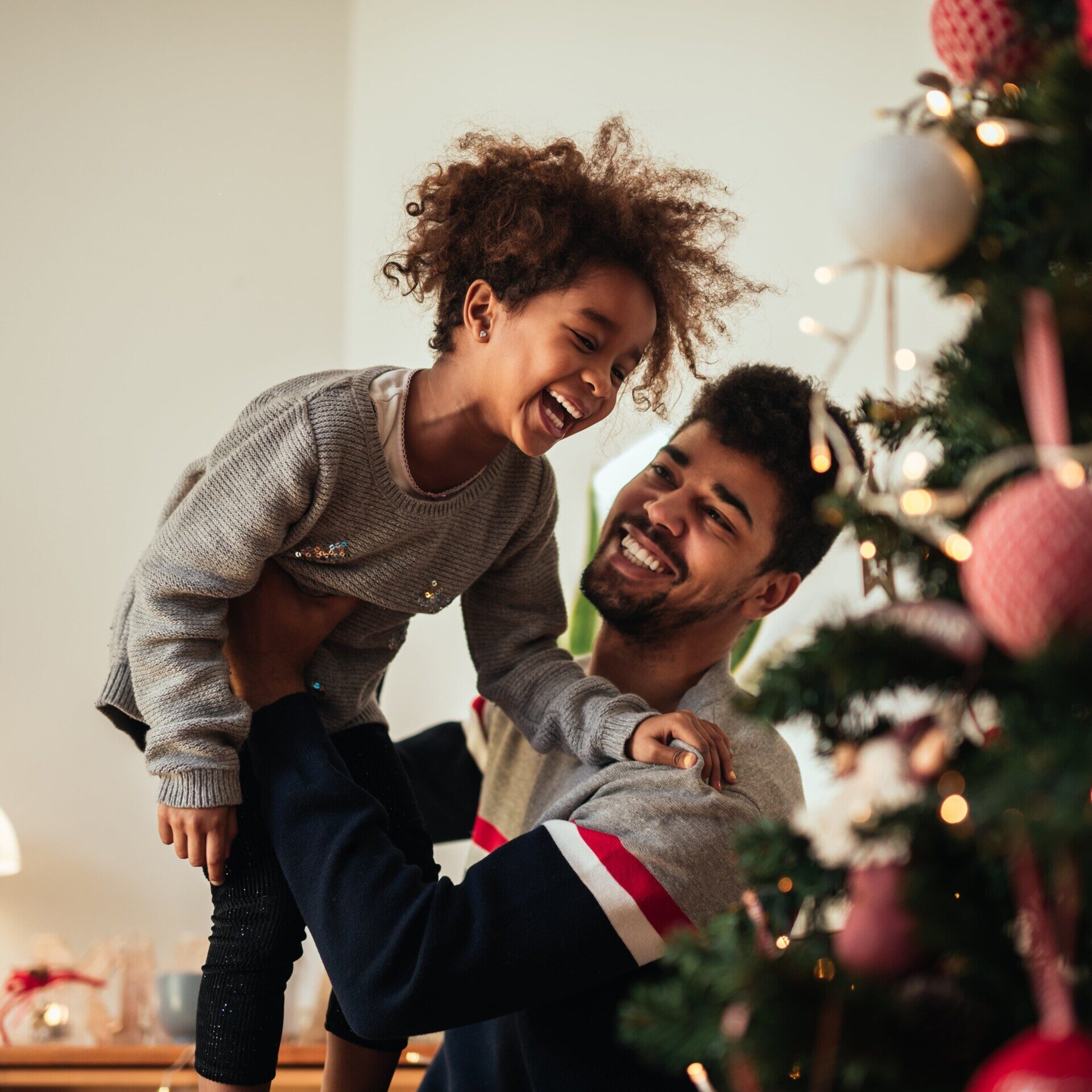 A man lifts a laughing child in front of a beautifully decorated Christmas tree, capturing the joy and warmth of the holiday season.